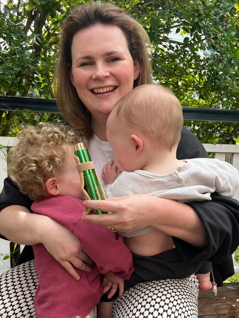 Woman is smiling holding a baby and toddler whose faces are away from the camera. She is holding an on-the-go stain spray called Shwink - in a glossy green aluminium bottle with a caramel colour stain-lifting cape. Green hedge and white palings in the background. 