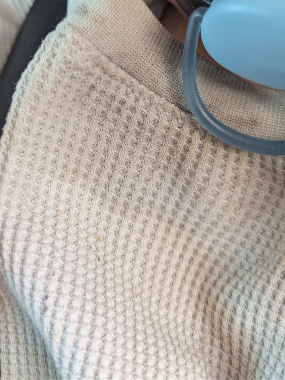 A close-up of a baby wearing a beige waffle-knit blanket and a black harness, with a light blue pacifier in their mouth.