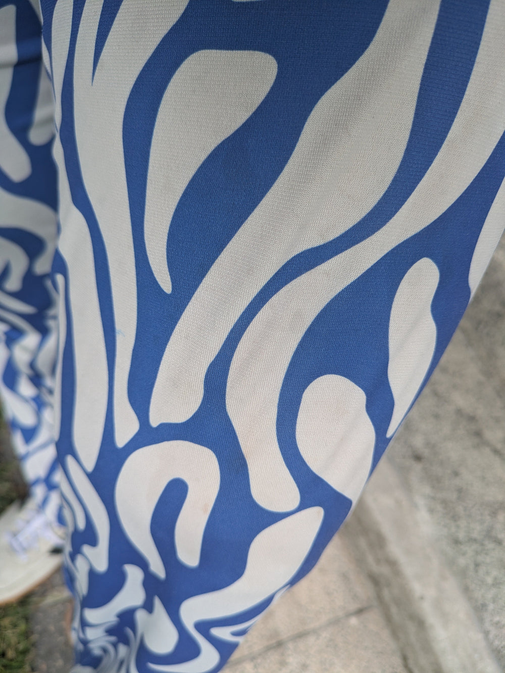 A close-up of patterned fabric with an abstract blue and off-white flame-like design, shown against a blurred outdoor ground background.