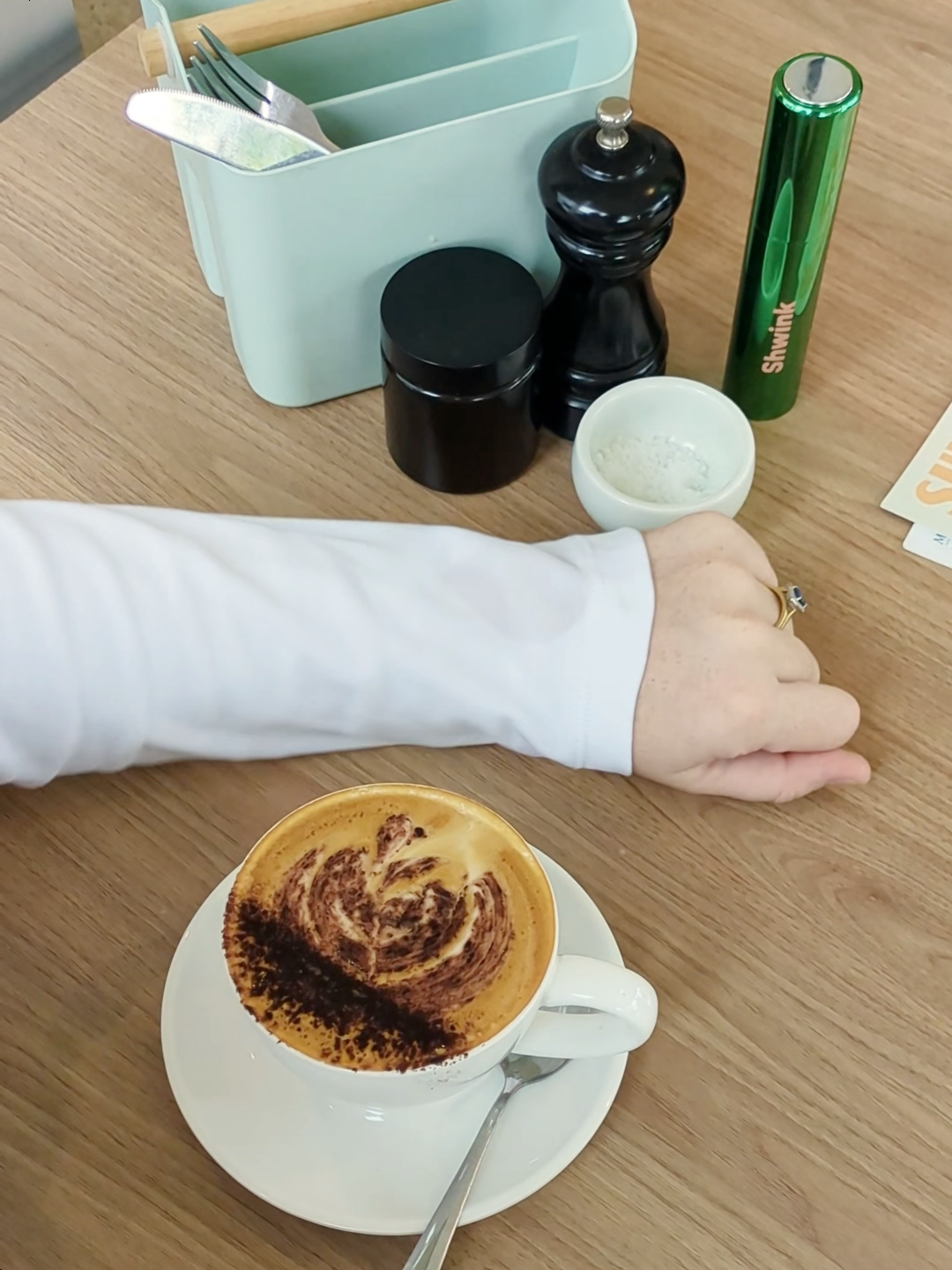 Cappuccino on a saucer with a hand reaching out, on a wooden table with coffee-making items. Stain on woman's white sleeve has been removed completely. Shwink on-the-go stain remover in glossy green aluminium bottle in the background next to black salt and pepper shakers. 