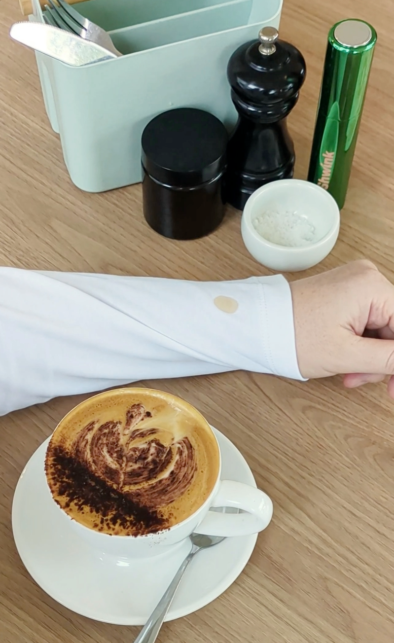 Cappuccino on a saucer with a hand reaching out, on a wooden table with coffee-making items. Stain on woman's white sleeve. Shwink on-the-go stain remover in glossy green aluminium bottle in the background next to black salt and pepper shakers. 