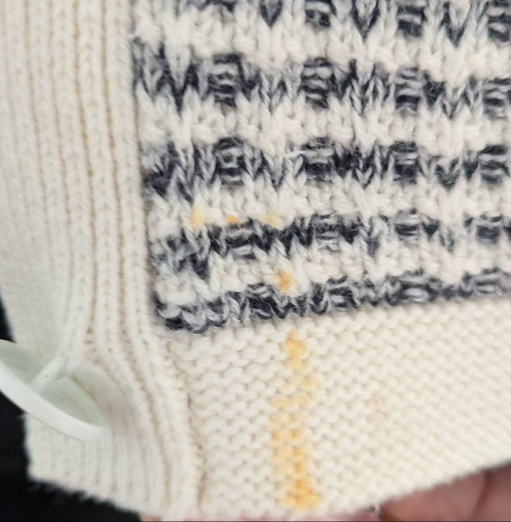 Close-up of a cream knit garment with a black and white patterned section, a white button, and a small yellow stain on the ribbed hem.