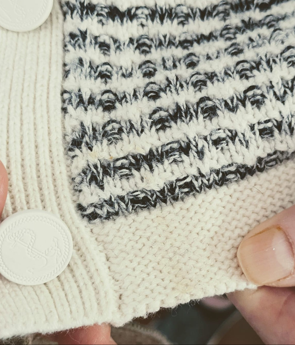 Close-up of a hand-knit cardigan featuring a cream ribbed trim with white buttons and a black-and-cream textured patterned body, held by hands.