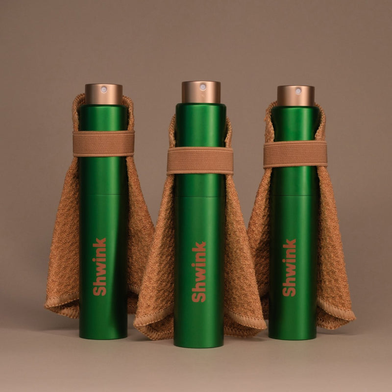 Three Shwink on-the-go stain spray bottles in matte with stain-lifting capes on a warm taupe background — travel stain remover spray.