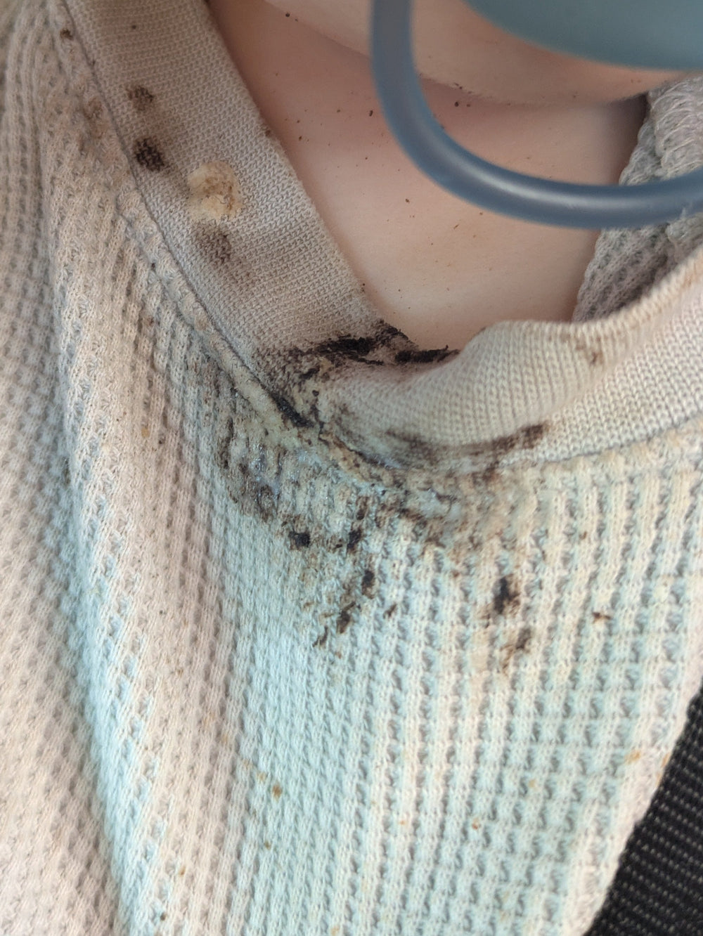A close-up of a baby wearing a dirty beige waffle-knit shirt and a black harness, with a blue pacifier in their mouth.