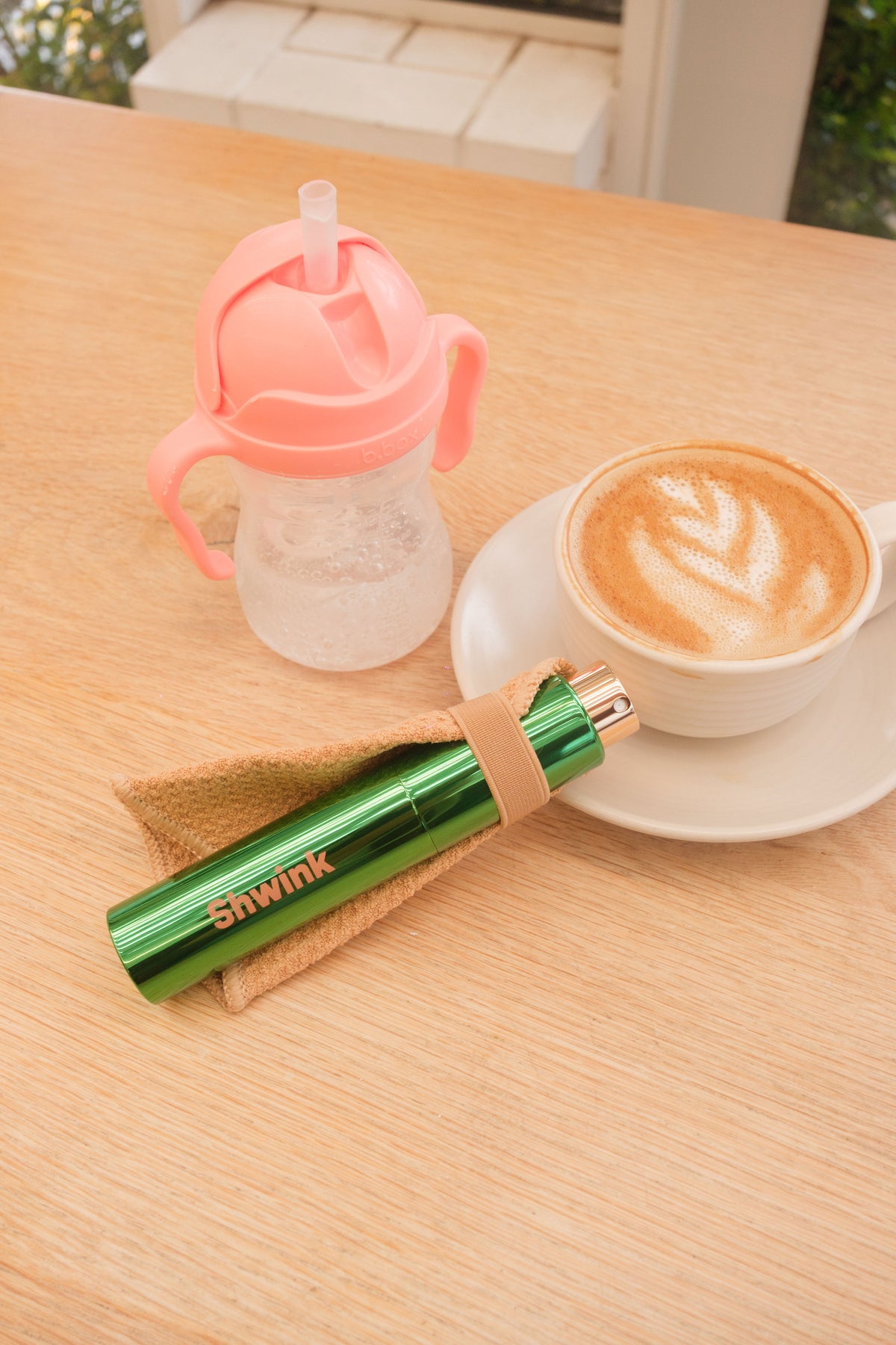 Resting on a light wood table are three items - a baby waterbottle with clear bottom and pink top, a flat white with coffee art in a white porcelain cup and saucer, and an on-the-go stain remover called Shwink in a glossy green aluminium bottle with a caramel coloured stain lifting cape. 
