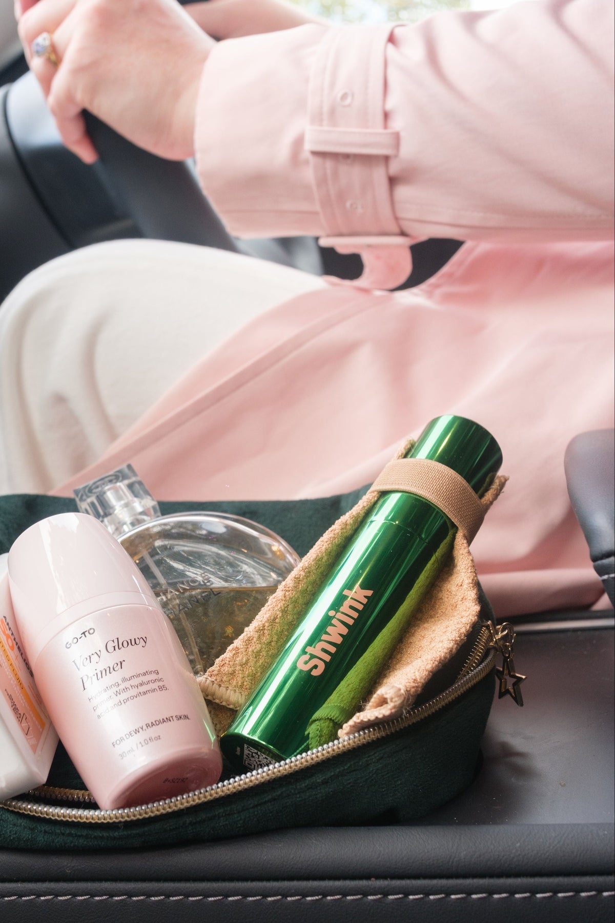 Woman sitting in Tesla car in light pink trench coat. Middle console shows with a green velvet makeup bag, a chanel perfume, a go-to skincare Primer and an on-the-go stain remover called Shwink in a glossy green aluminium bottle with a stain lifting cape in a caramel colour. 