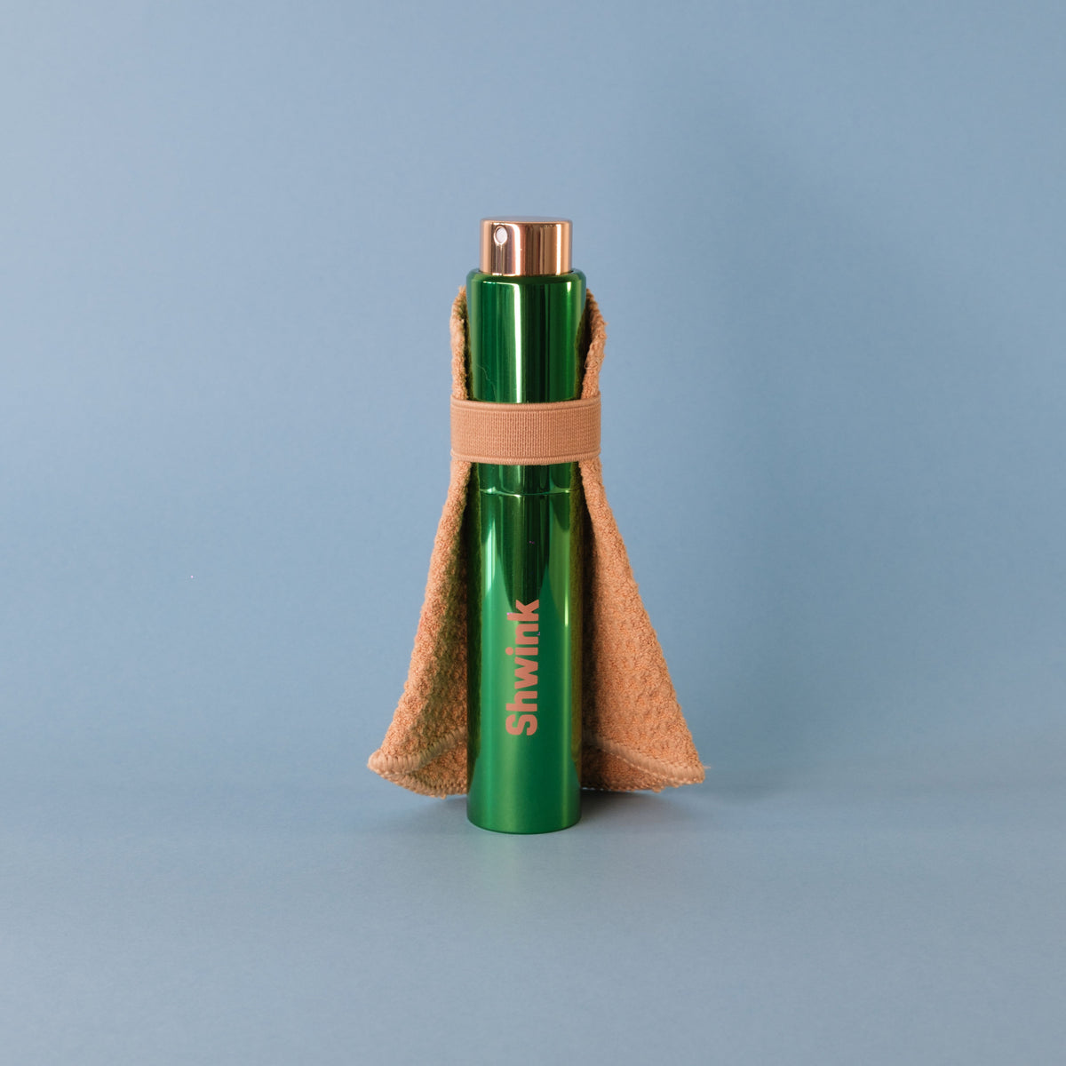 A green cylindrical bottle with a metallic rose gold cap, a Shwink on-the-go stain spray with stain-lifting cape, standing on a light blue-gray background.