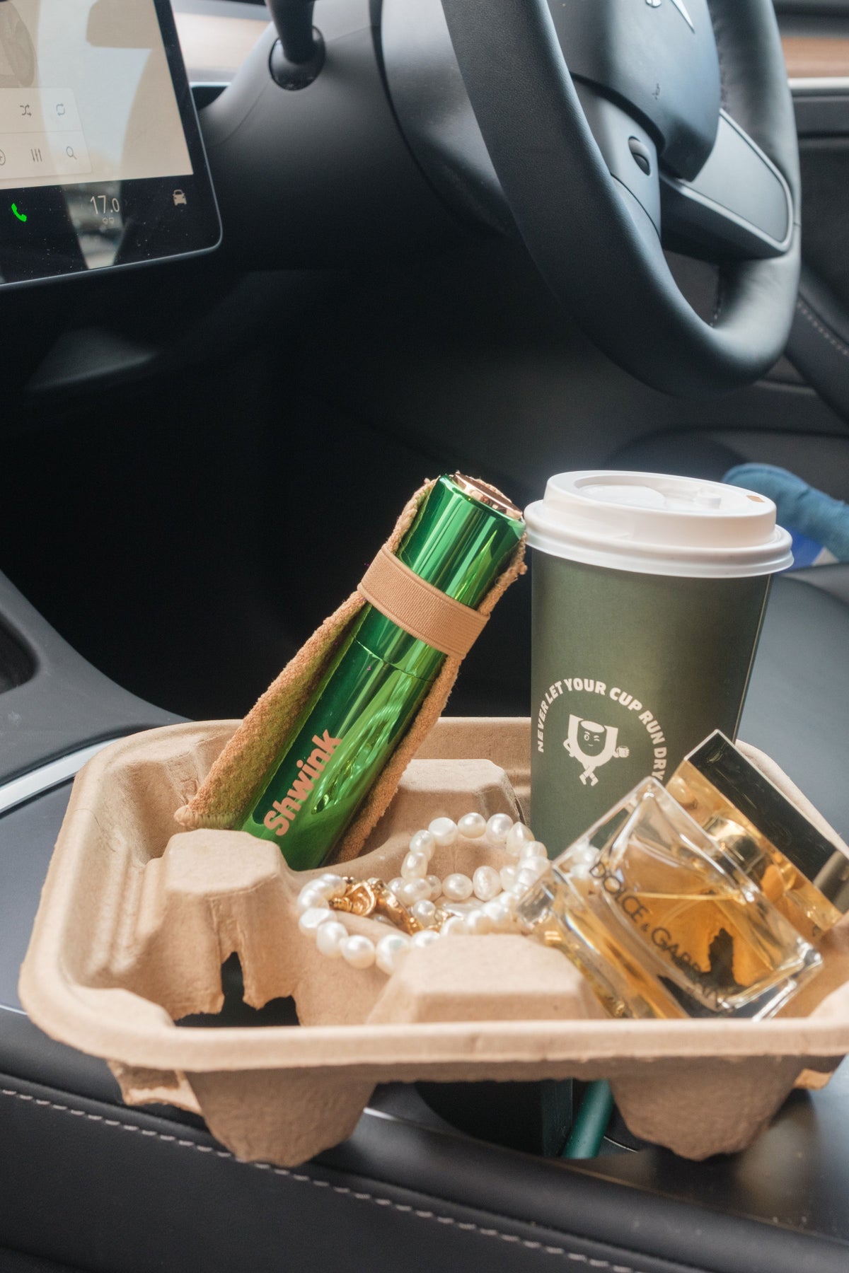 A coffee cup, a Shwink on-the-go stain spray with stain-lifting cape (glossy green aluminium cylindrical tube with caramel stain lifting cape), a pearl necklace, and a small perfume bottle resting in a brown paper tray on a car center console.