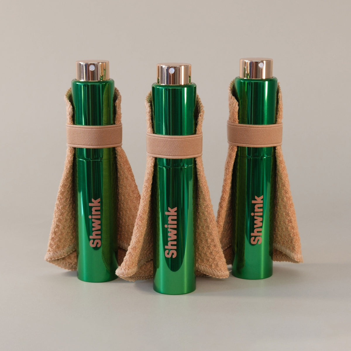 Three green cylindrical Shwink bottles wrapped in stain lifting cape standing on a light gray surface against a neutral light gray background.