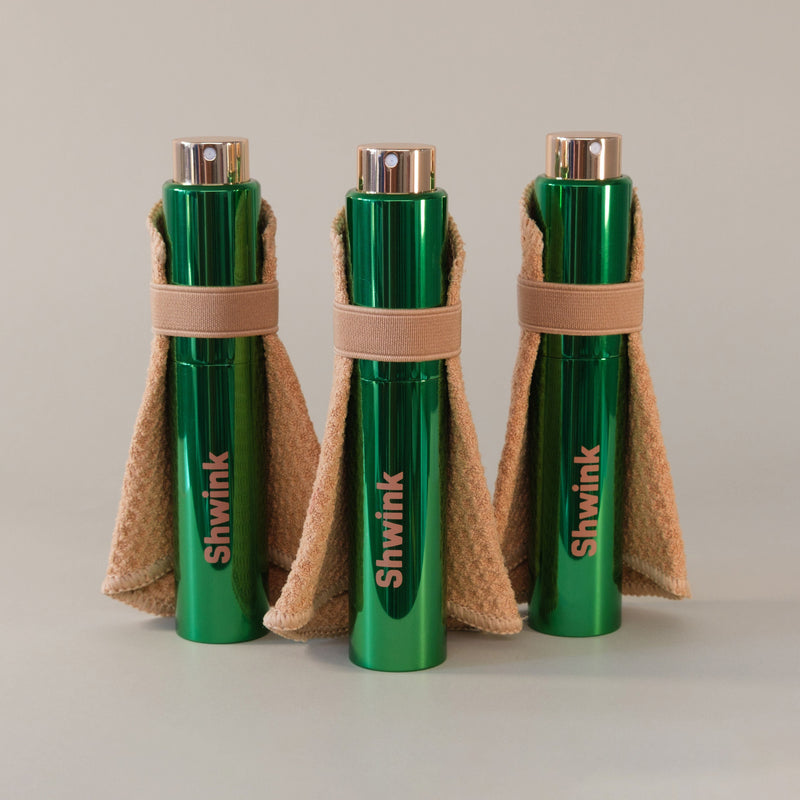 Three green cylindrical Shwink bottles wrapped in stain lifting cape standing on a light gray surface against a neutral light gray background.