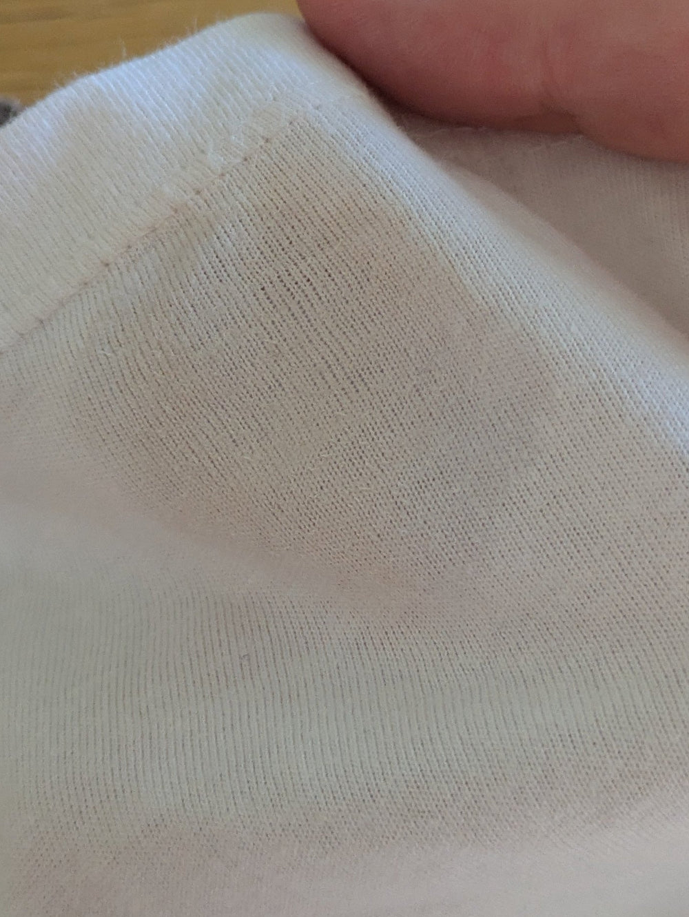 A close-up of a hand holding a folded cream-colored cotton muslin fabric, with a brown textured baby wipe pad underneath and a blue packet of wipes on a wooden table to the left.
