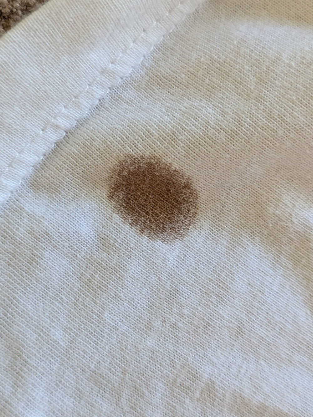 A close-up of a white fabric item, likely a diaper or baby blanket, showing a brown stain on the front. The fabric has a stitched border and is laid on a brown carpet.